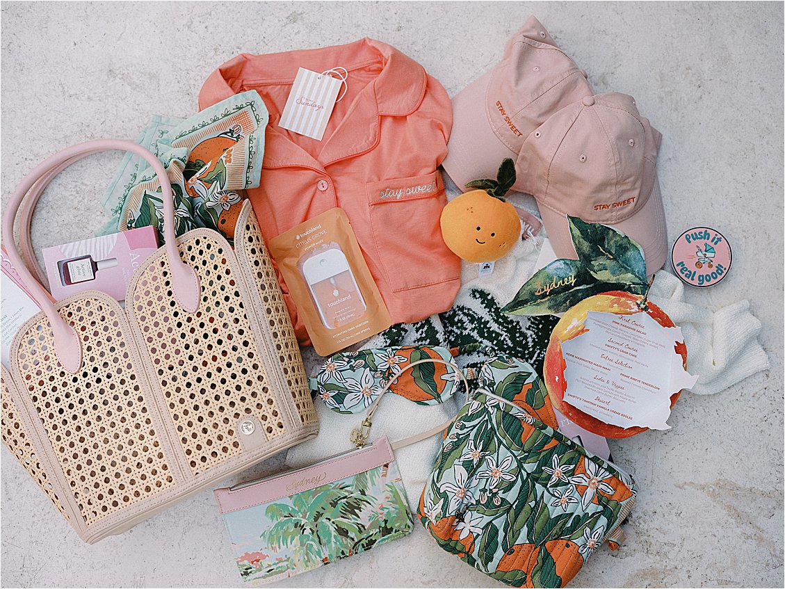 Flat lay of Zoe Strollers corporate retreat welcome bag contents including salmon pajamas, pink baseball cap, orange plush toy, rattan tote, botanical scarf, and personalized itinerary, styled by Gramercy & Grace at The Colony Hotel Palm Beach