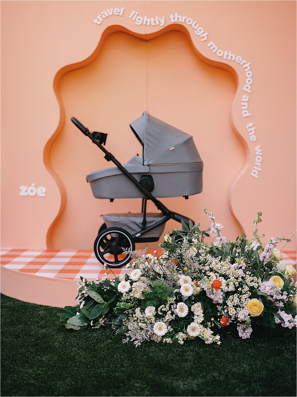 Zoe stroller displayed in a custom peach wavy arch backdrop reading "travel lightly through motherhood and the world," surrounded by a lush floral ground arrangement at The Colony Hotel Palm Beach corporate retreat 