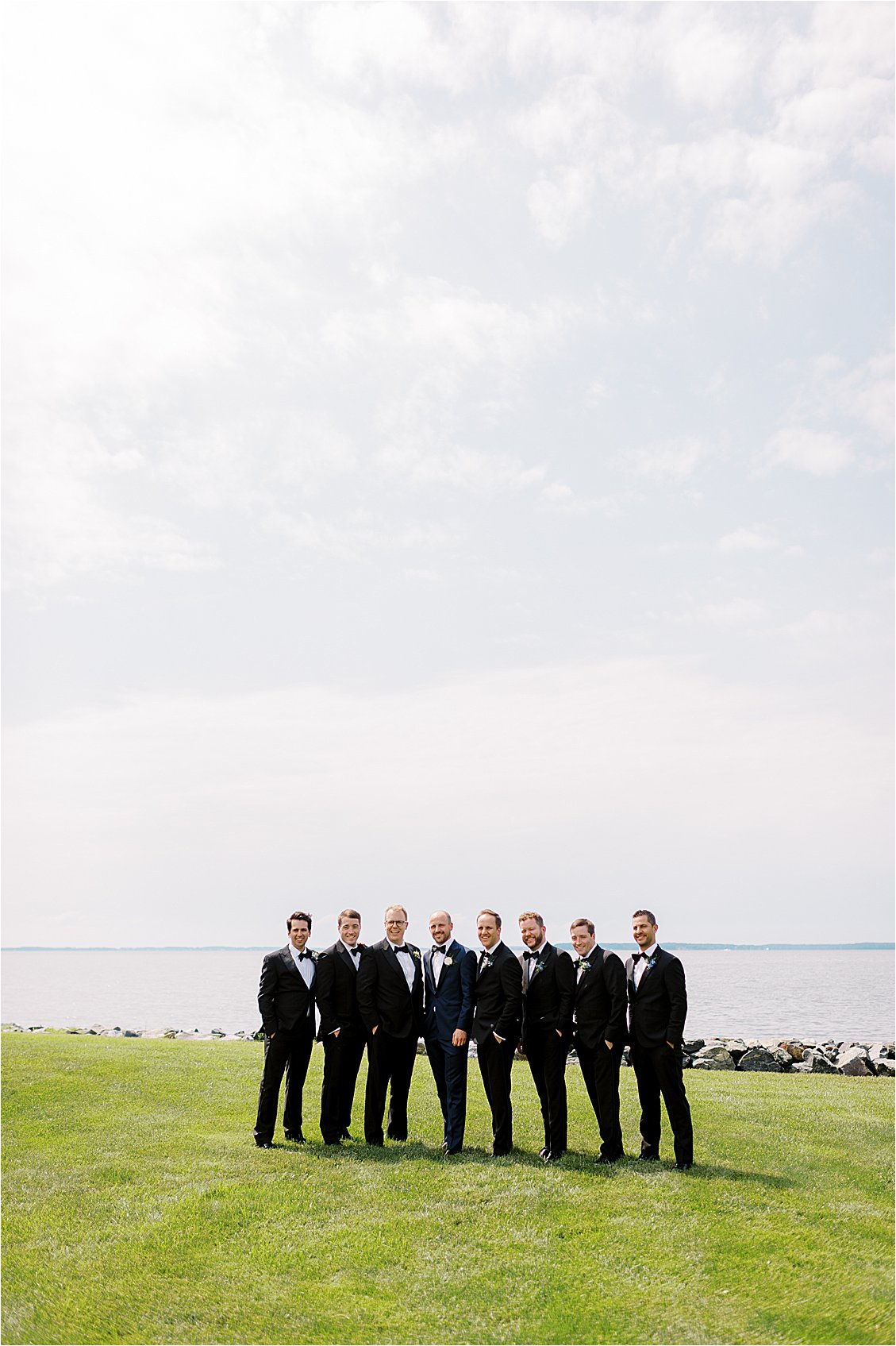 Groom and groomsmen in black tuxedos posing along the Chesapeake Bay shoreline at a private waterfront estate wedding in Oxford Maryland.