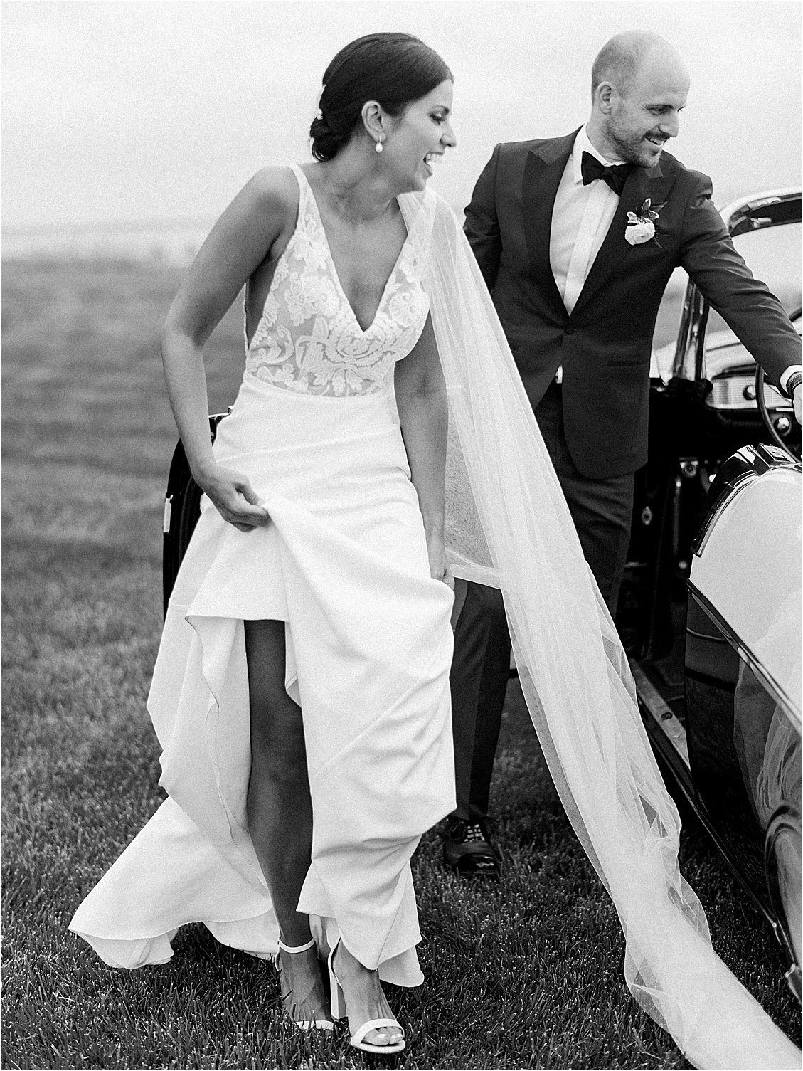 Black and white wedding portrait of bride and groom stepping out of vintage convertible at waterfront estate