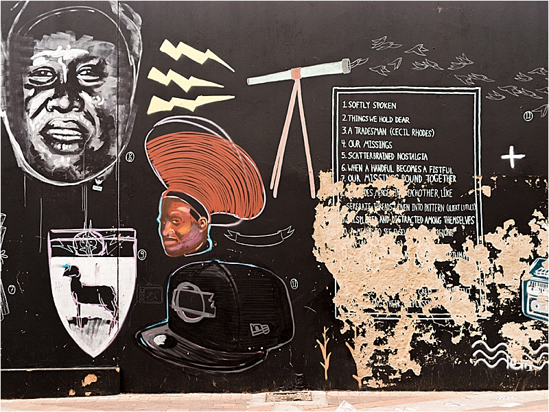 Bold street art mural on a black wall in Maboneng Precinct, Johannesburg, featuring painted portraits and handwritten text, photographed during a Johannesburg South Africa travel guide visit by Renee Hollingshead