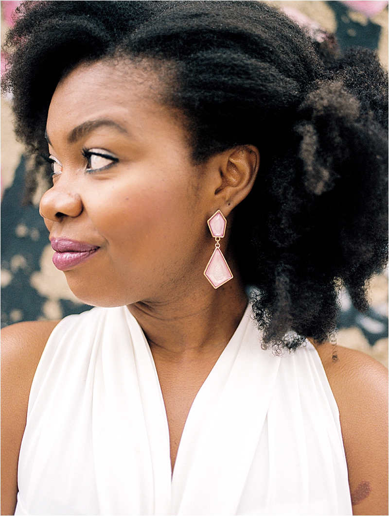 Close-up film portrait of Aisha of Aisha and Life wearing a white halter dress and pink geometric earrings, looking off to the side in front of a floral mural at Maboneng Precinct in Johannesburg, South Africa, photographed by Renee Hollingshead