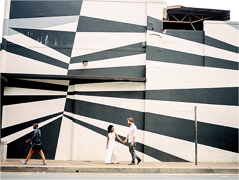 Film image of Aisha and Kev walking hand in hand in front of a large black and white geometric mural on a building in Maboneng Precinct, Johannesburg, South Africa, during their anniversary session with destination film photographer Renee Hollingshead
