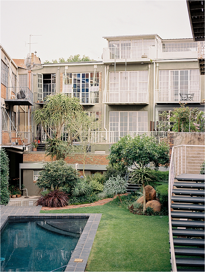 Film image of a lush Johannesburg Airbnb courtyard featuring a dark pool, manicured lawn, tropical garden, and multi-story building with white balconies, photographed by destination film photographer Renee Hollingshead
