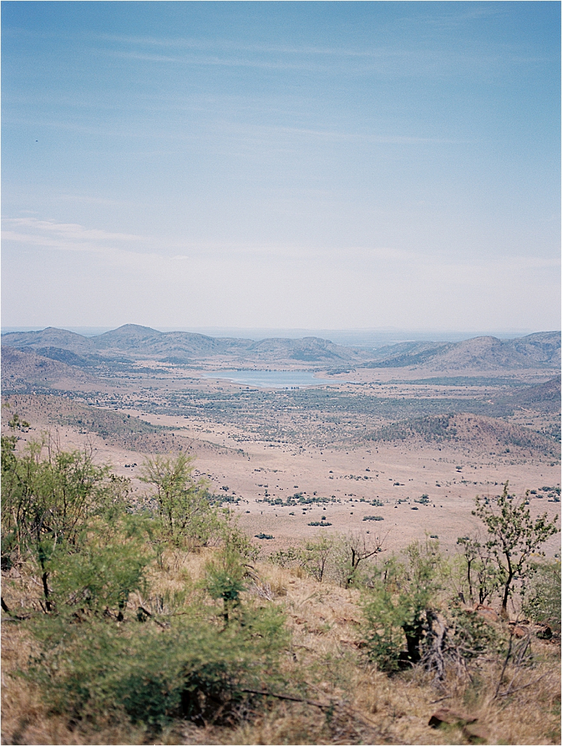 Expansive film landscape of Pilanesberg National Park, South Africa, showing dry golden savanna, sparse green shrubs, rolling mountains, and a distant lake under a hazy blue sky, photographed by destination film photographer Renee Hollingshead