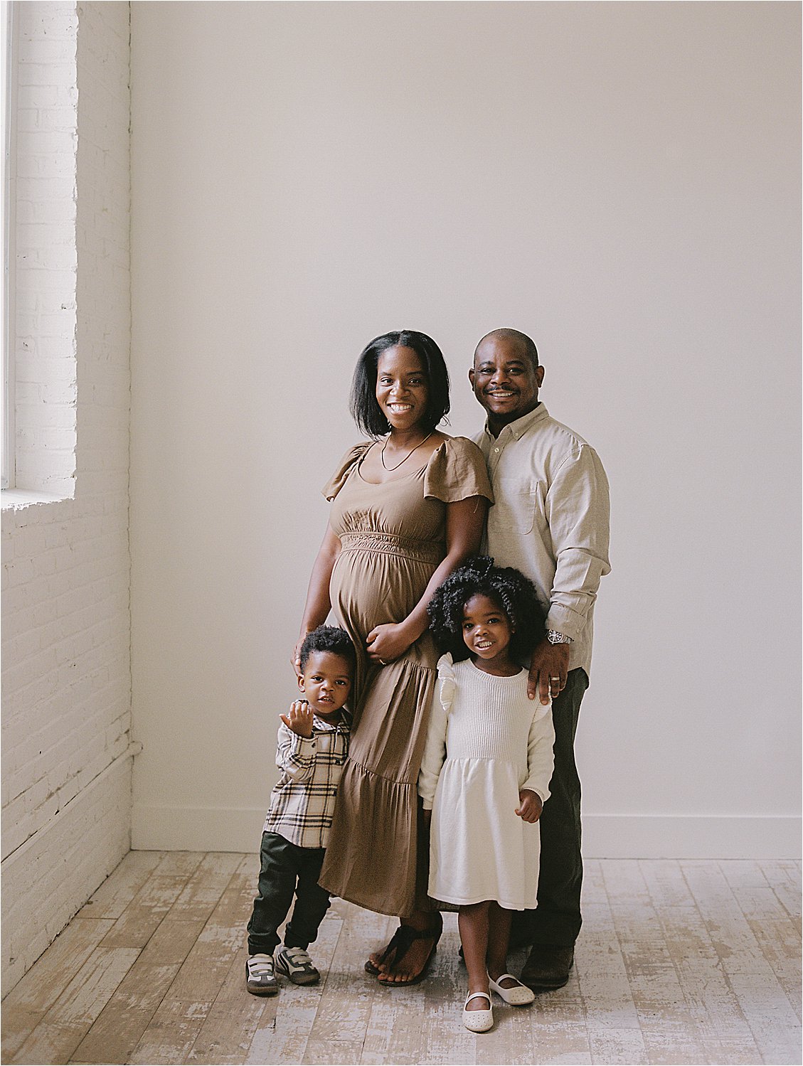 Family of four posing near studio window during film maternity session in Annapolis Maryland, expecting mother in camel dress, photographed by Renee Hollingshead Photography