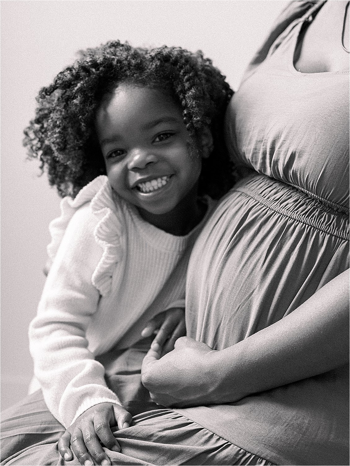 Black and white close up of young girl smiling while hugging expecting mother's baby bump, studio maternity session Annapolis Maryland by Renee Hollingshead Photography