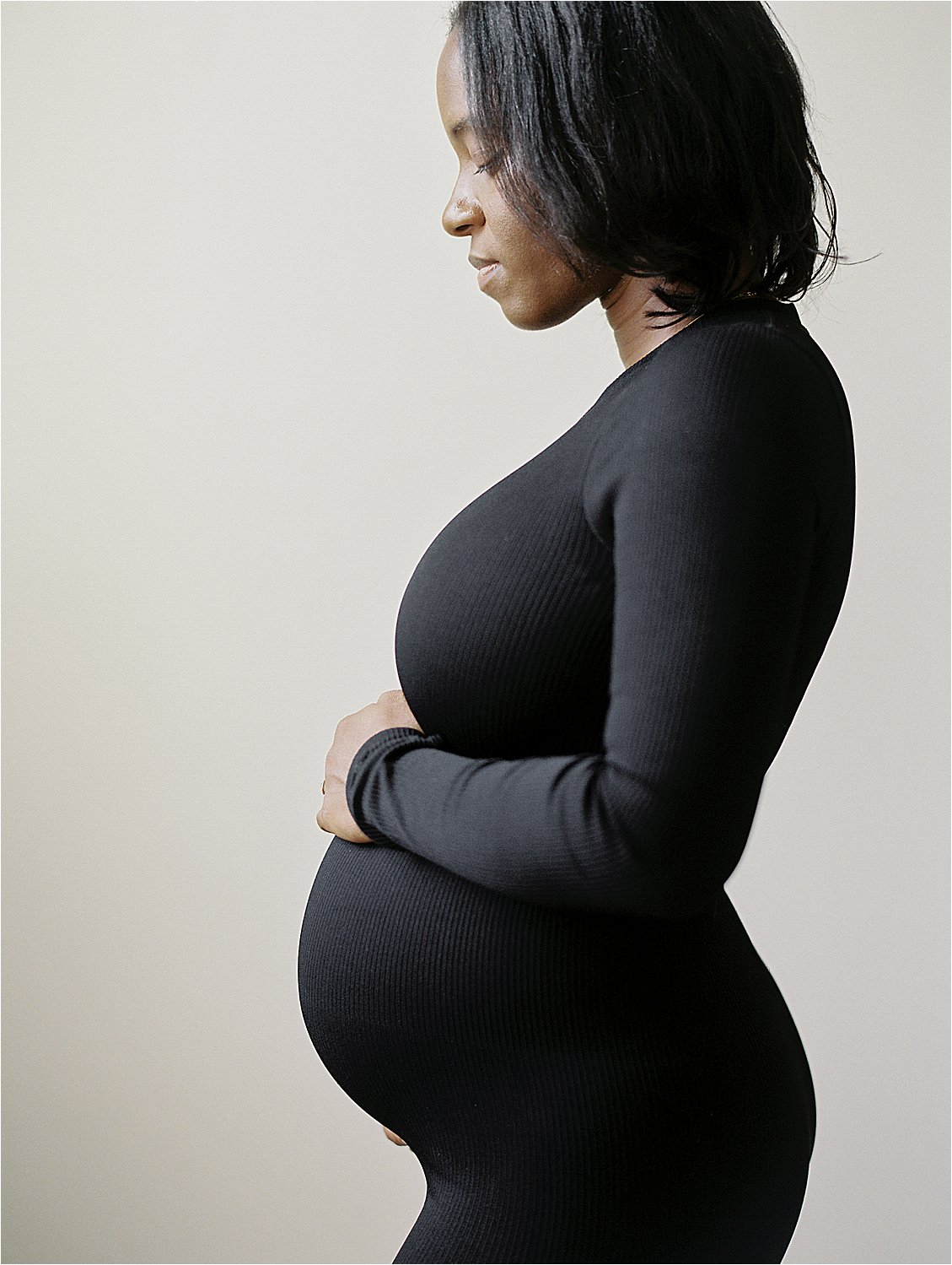 Side profile of expecting mother in black turtleneck dress cradling baby bump in studio, film maternity portrait Annapolis Maryland by Renee Hollingshead Photography