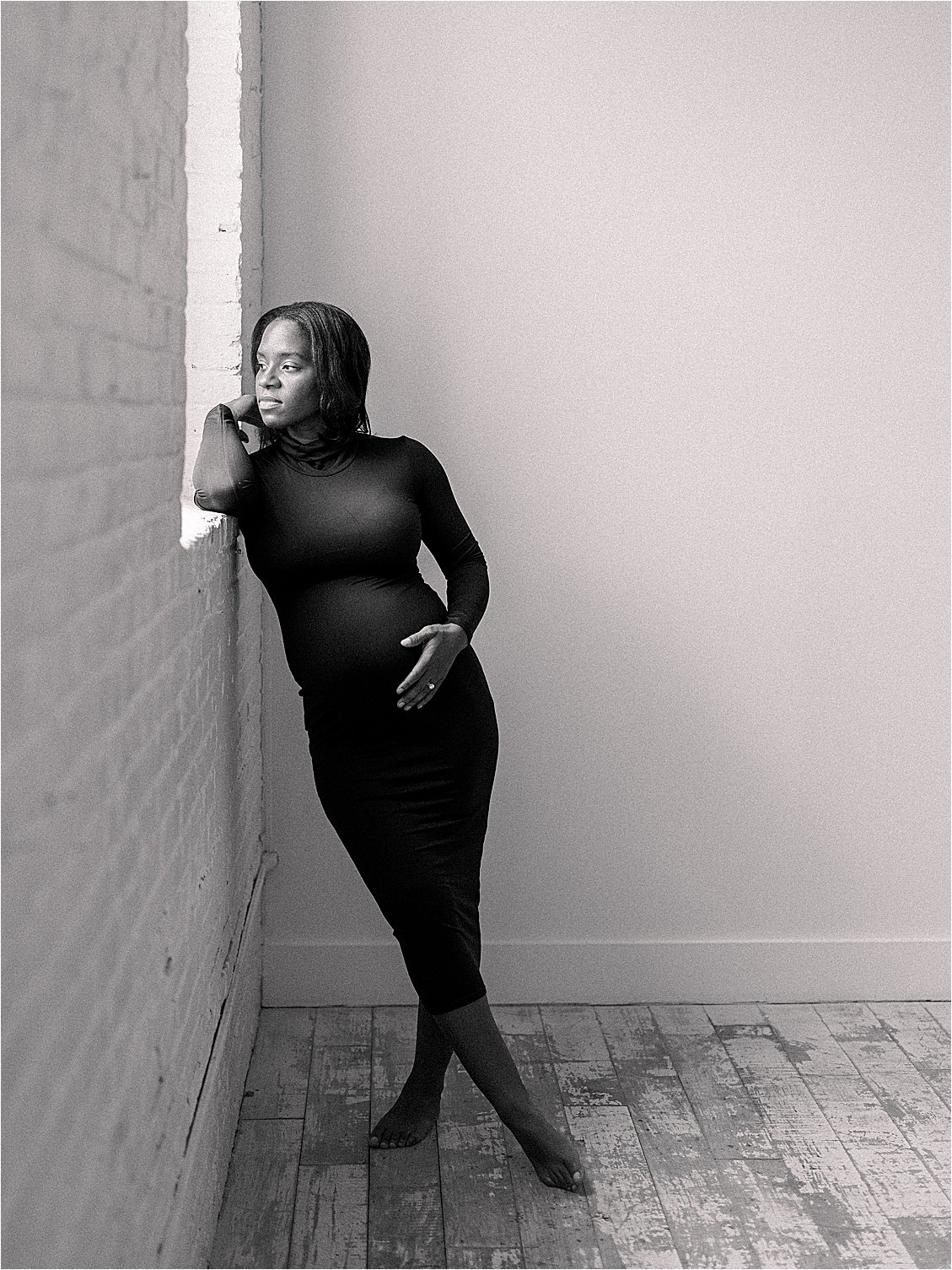 Black and white editorial portrait of expecting mother in black dress leaning against white brick wall, studio maternity session Annapolis Maryland by Renee Hollingshead Photography