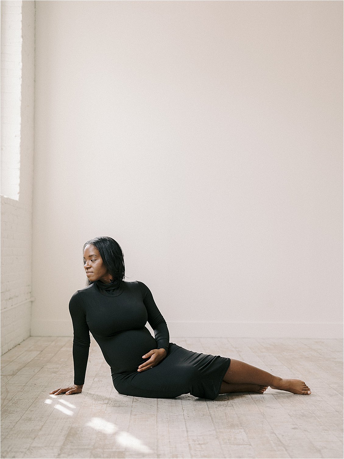 Expecting mother in black turtleneck dress seated on studio floor in a beam of light, cradling baby bump, film maternity session Annapolis Maryland by Renee Hollingshead Photography