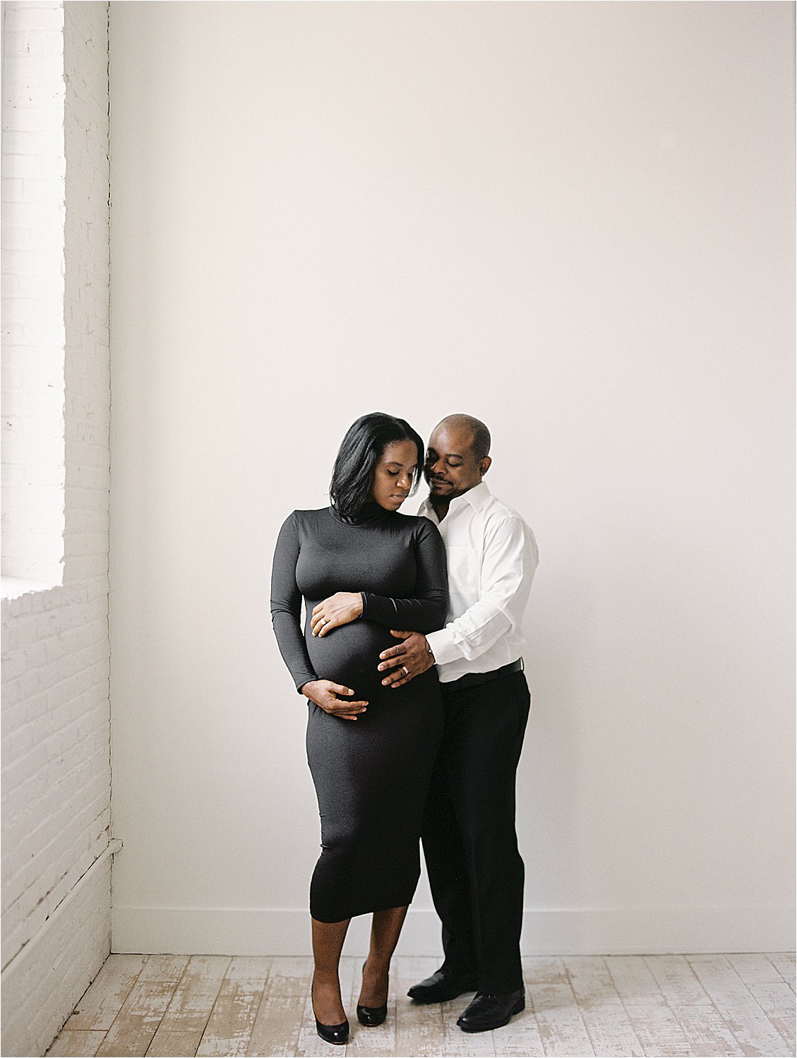 Intimate studio portrait of expecting couple in black, husband resting chin on wife's shoulder as both cradle baby bump, film maternity session Annapolis Maryland by Renee Hollingshead Photography