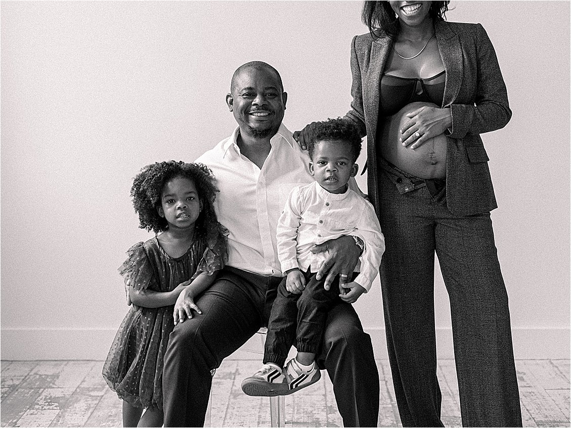 Black and white studio family portrait with father smiling at camera, two children on his lap, and expecting mother standing beside them with exposed bump, film maternity session Annapolis Maryland by Renee Hollingshead Photography