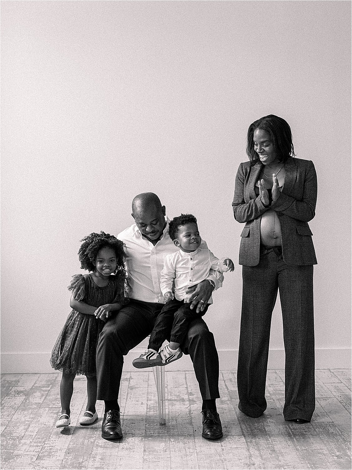 Candid black and white studio portrait of family laughing together — father and two kids seated, expecting mother standing beside them in blazer, film maternity session Annapolis Maryland by Renee Hollingshead Photography