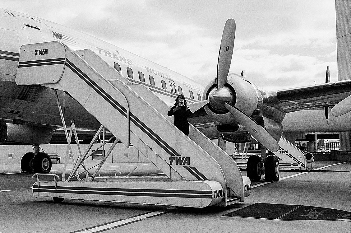 Photographer Renee Hollingshead holding a film camera on the stairs of a vintage TWA airplane, black and white film photography