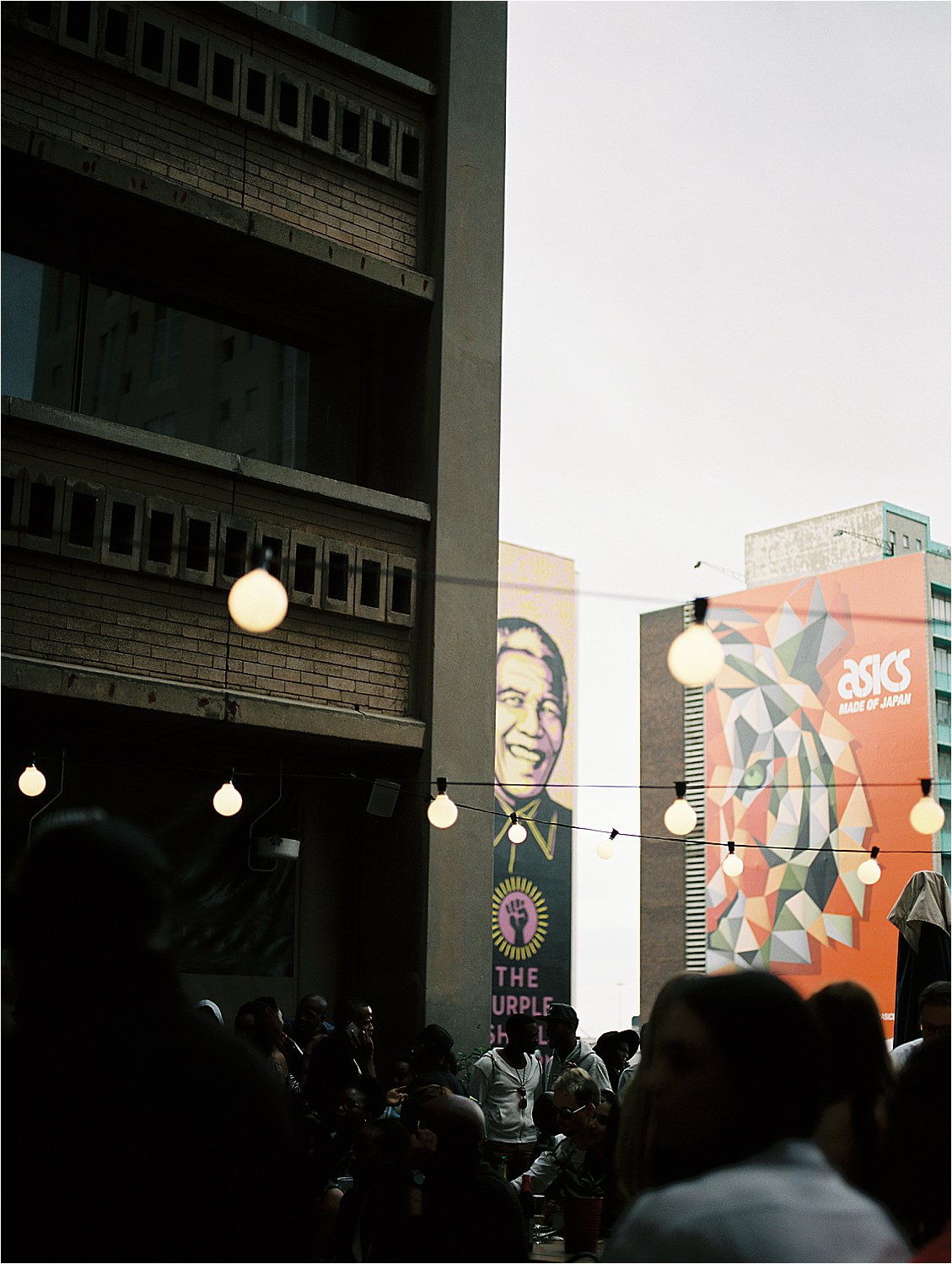 Image 1 (single — string lights + murals) Evening atmosphere at Neighbourgoods Market in Johannesburg, South Africa — warm globe string lights hang between buildings as a crowd gathers below, with large colorful murals including a Nelson Mandela portrait visible on surrounding city buildings, photographed on film by destination film photographer Renee Hollingshead