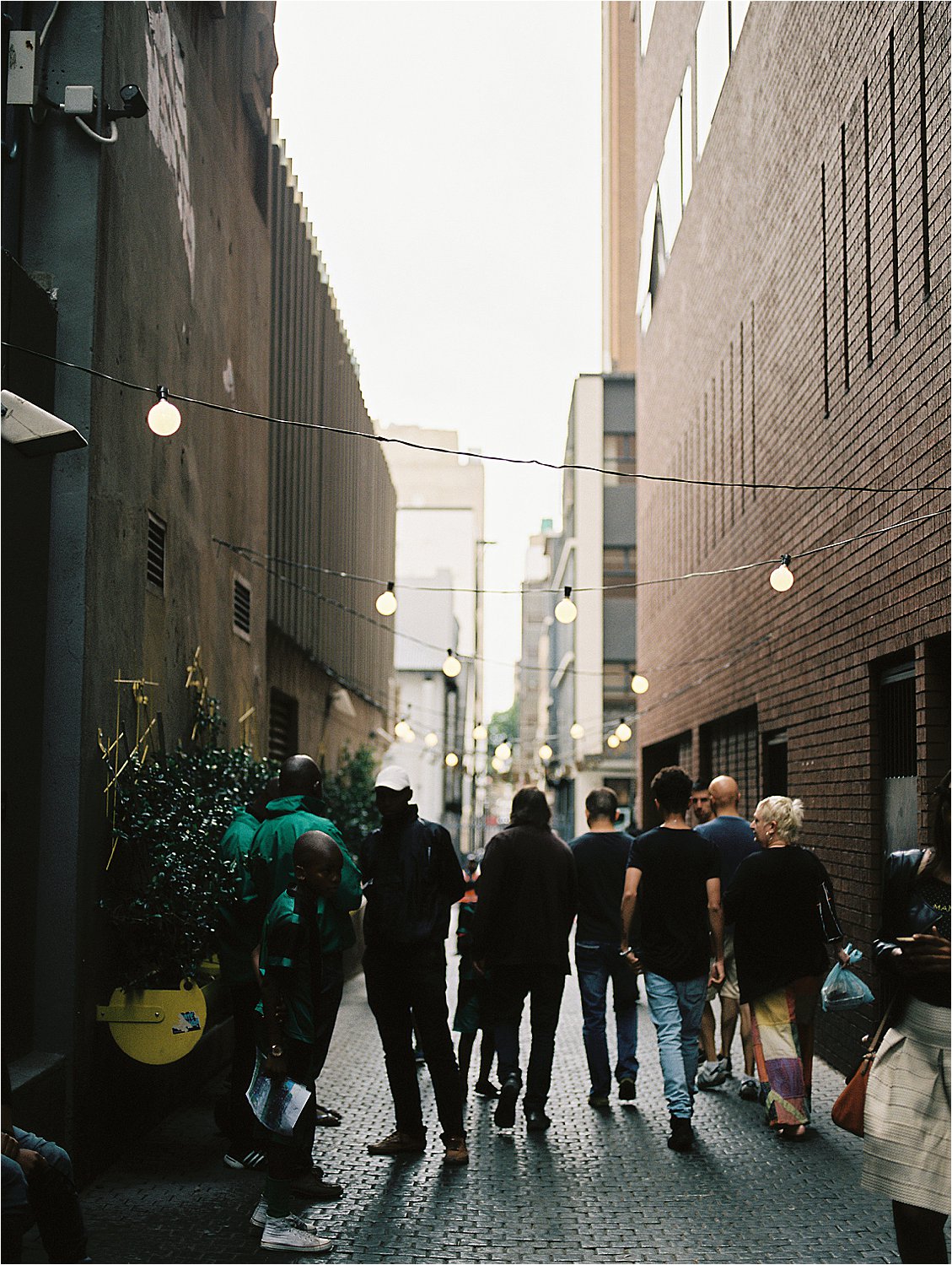 Film image of a narrow brick alley at Neighbourgoods Market in Johannesburg, South Africa, lined with warm globe string lights strung overhead and a diverse crowd of people walking together toward the market entrance, photographed by destination film photographer Renee Hollingshead