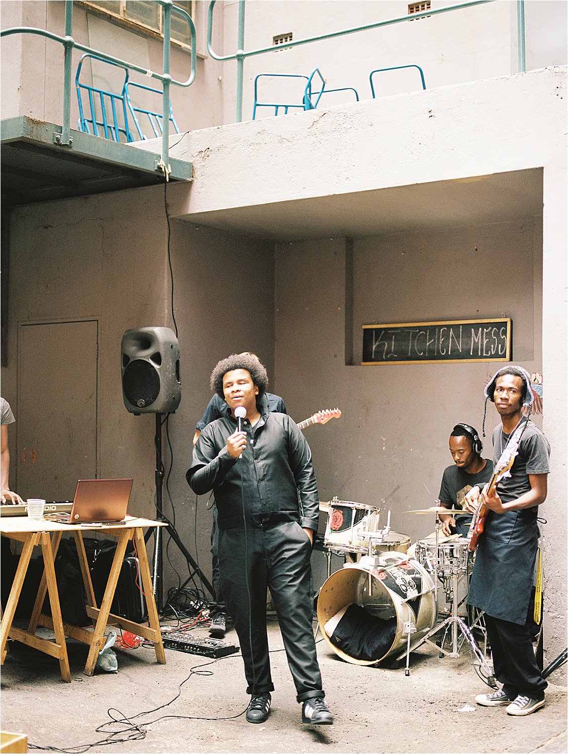 Live band performing outdoors at Neighbourgoods Market in Johannesburg, South Africa on film — a vocalist in all black holds a microphone center stage while a drummer and guitarist perform behind him beneath a chalkboard sign reading "Kitchen Mess," photographed by destination film photographer Renee Hollingshead
