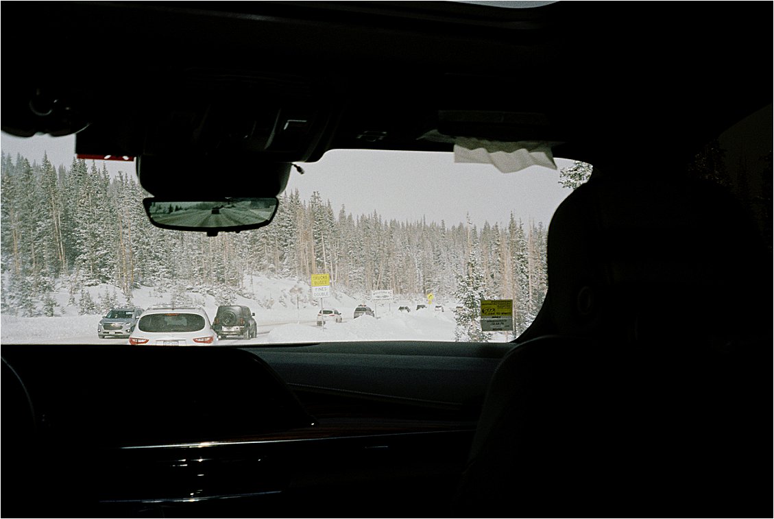 Snow-covered highway lined with pine trees on the drive to Winter Park Colorado ski resort, shot from inside a car on film. Film travel photography by Renee Hollingshead on Kodak ColorPlus 200.