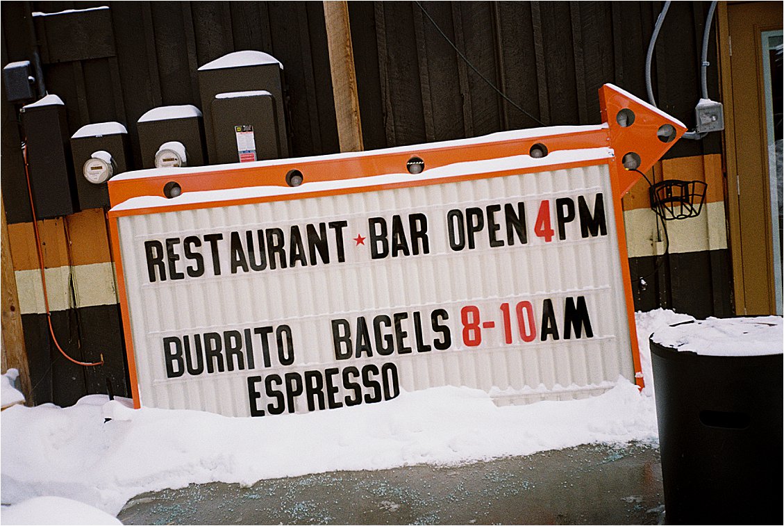 Retro orange marquee sign for a Winter Park Colorado restaurant and bar reading "Burrito Bagels 8–10AM, Espresso" dusted with snow. Film photo by destination photographer Renee Hollingshead on Kodak ColorPlus 200. 