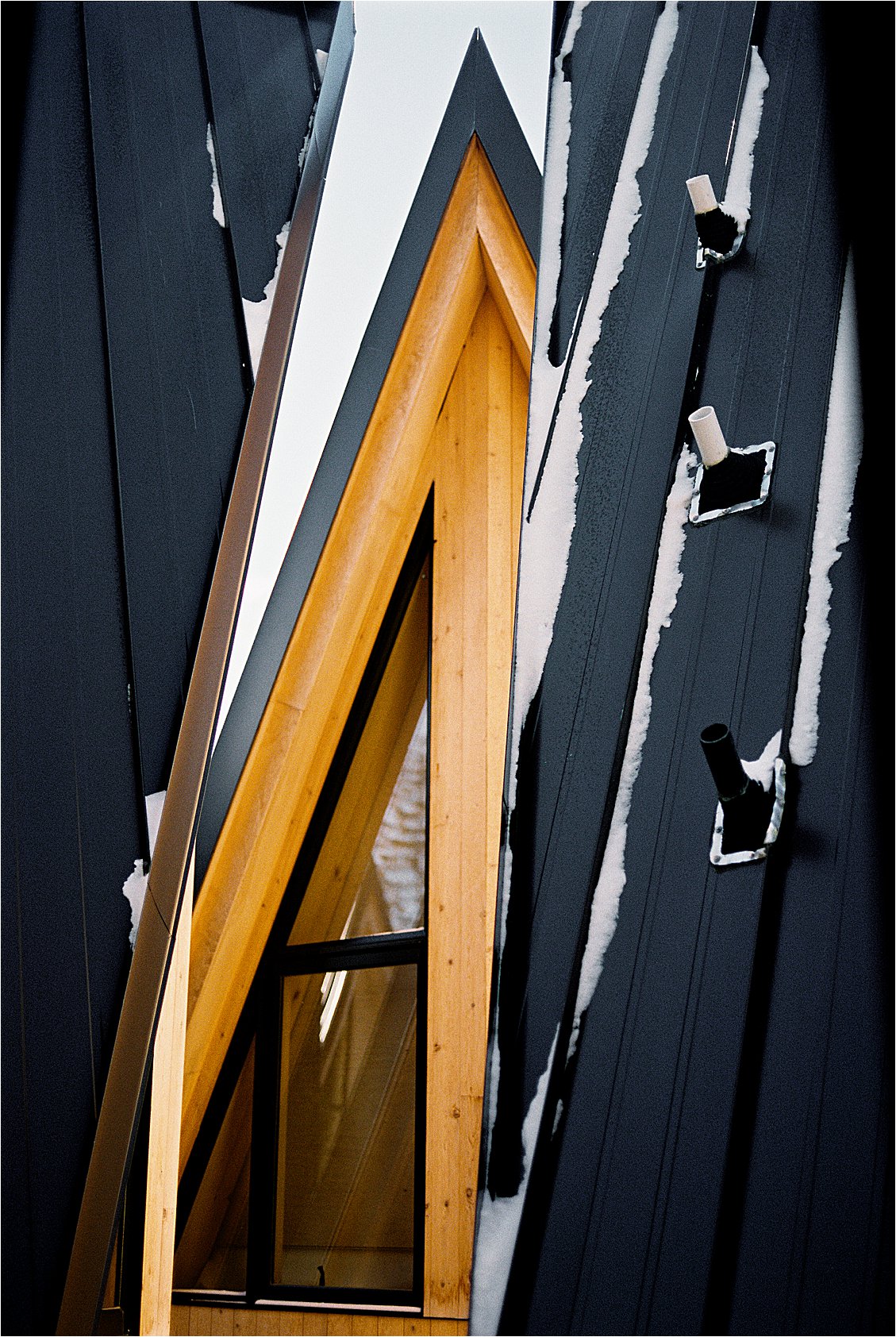 Close-up architectural detail of the A-frame roofline at A-Frame Club hotel in Winter Park Colorado — warm pine wood beams and dark metal panels dusted with fresh snow. Film photo by Renee Hollingshead on Kodak Portra 400. 