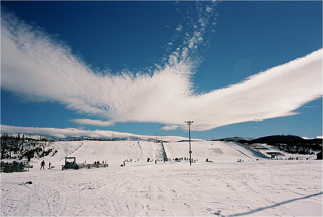 Wide open ski slopes at Winter Park Resort Colorado under a vivid blue sky with dramatic wispy clouds, skiers visible in the distance on a sunny winter day. Film photo by Renee Hollingshead on Kodak ColorPlus 200.