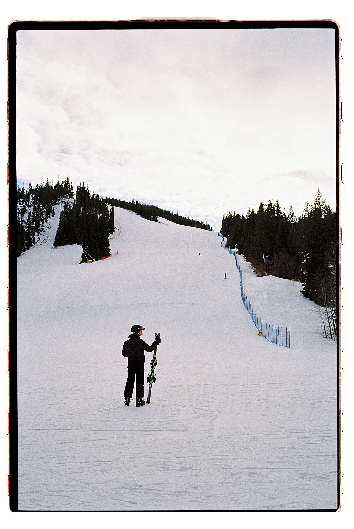 Lone skier standing at the base of a wide groomed run at Winter Park Resort Colorado, skis in hand, pine-covered slopes stretching into the distance. Film photo by Renee Hollingshead on Kodak Portra 400. 