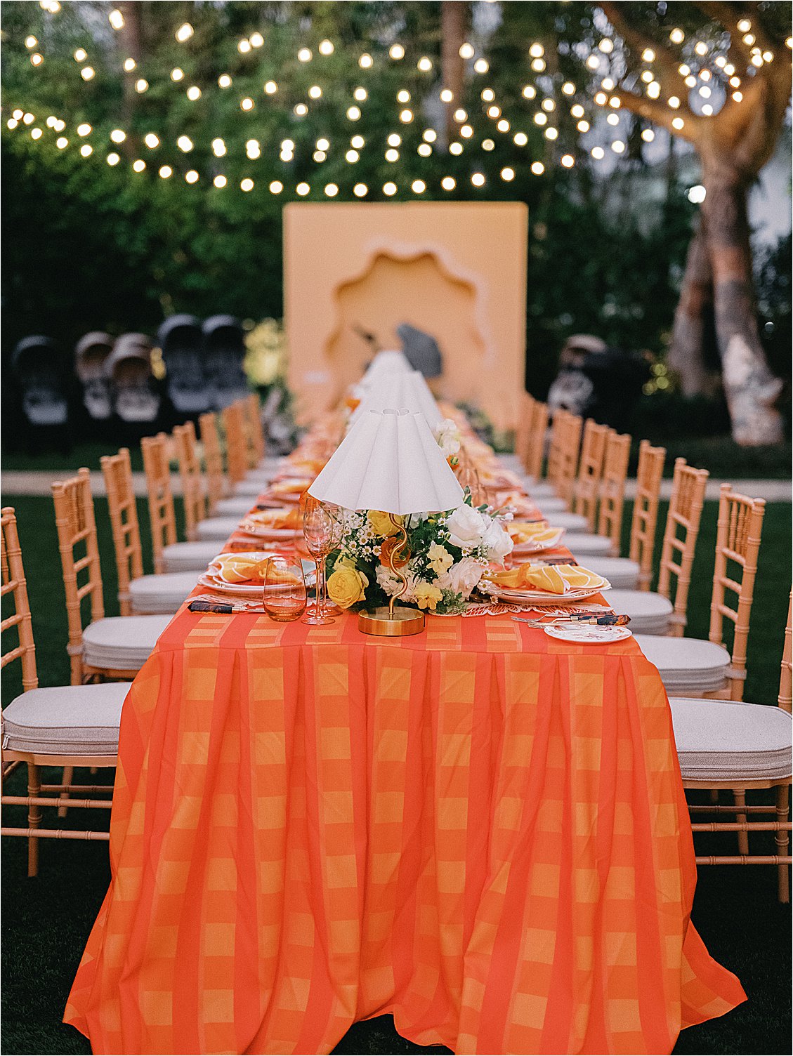 Straight-on view down the entire long corporate dinner table with orange plaid tablecloth, floral centerpieces, and white table lamps leading to the Zoe brand backdrop, under café string lights at The Colony Hotel Palm Beach East Garden 