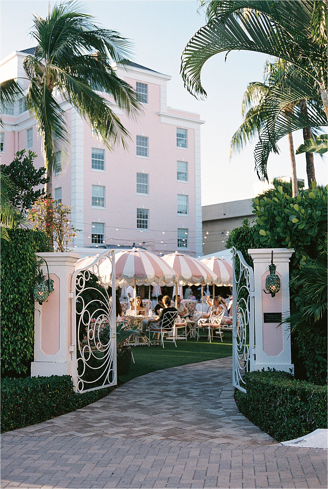 The Colony Hotel Palm Beach ornate white iron East Garden gate open to an outdoor gathering under pink and white striped umbrellas with string lights and the blush pink tower in the background, photographed on film