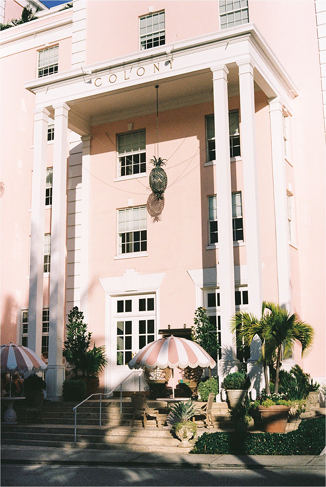 The Colony Hotel Palm Beach blush pink front entrance with white columns, hanging pineapple chandelier, and striped umbrellas on the front steps, photographed on film