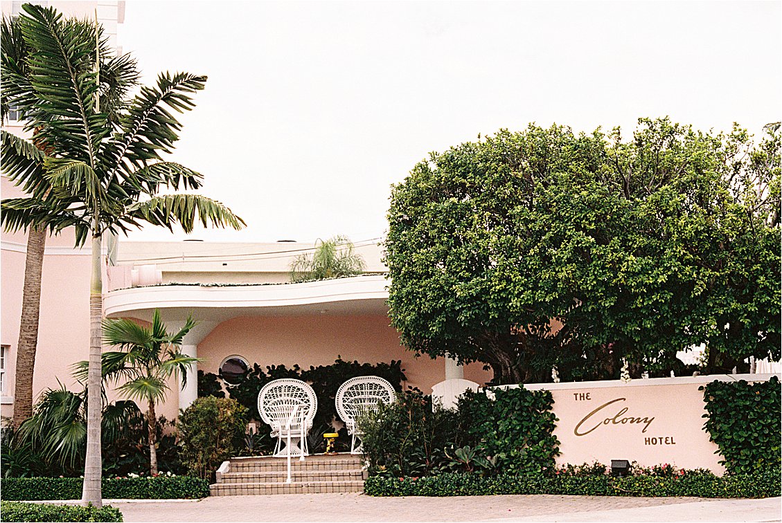 The Colony Hotel Palm Beach Entrance Sign and Peacock Chairs Alt Text: The Colony Hotel Palm Beach blush pink facade with white peacock chairs and hotel sign surrounded by tropical landscaping, photographed on film