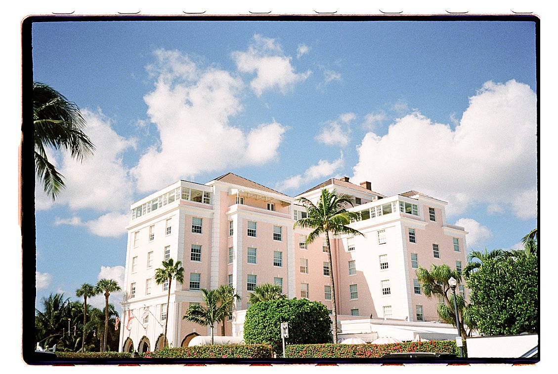 Wide film photograph of The Colony Hotel Palm Beach full blush pink exterior with white trim, palm trees, and dramatic blue sky with white clouds