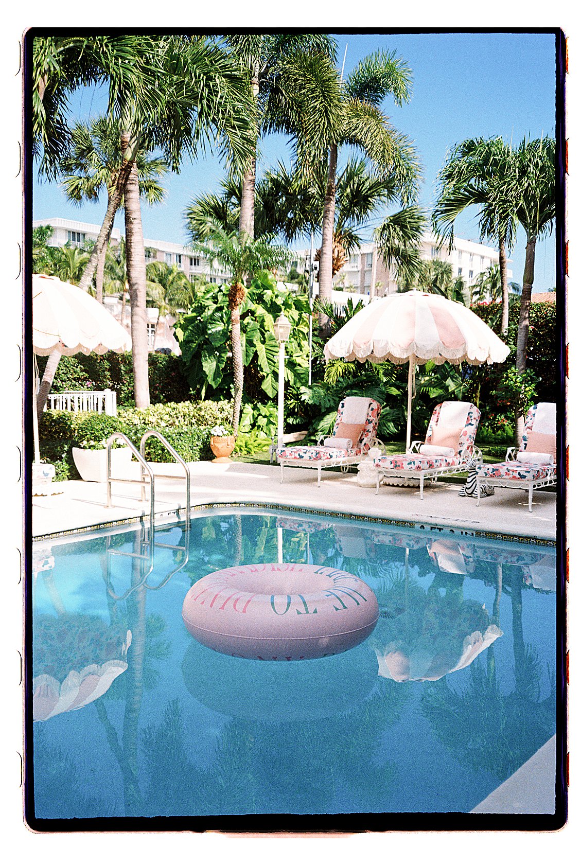 The Colony Hotel Palm Beach pool with a pink pool float, fringed umbrella, floral chaise lounges, and lush tropical garden backdrop, photographed on film