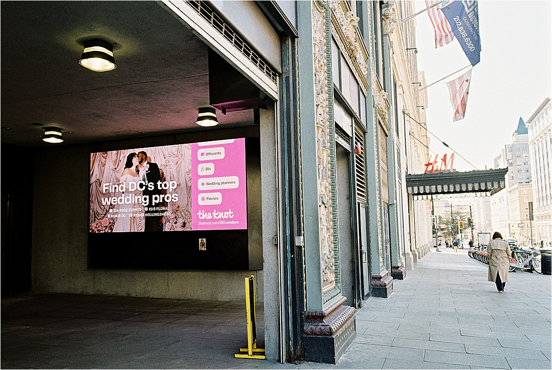 The Knot Washington DC wedding photographer billboard displayed on a building exterior in Penn Quarter, with the DC flag and ornate historic architecture visible on the street alongside it.