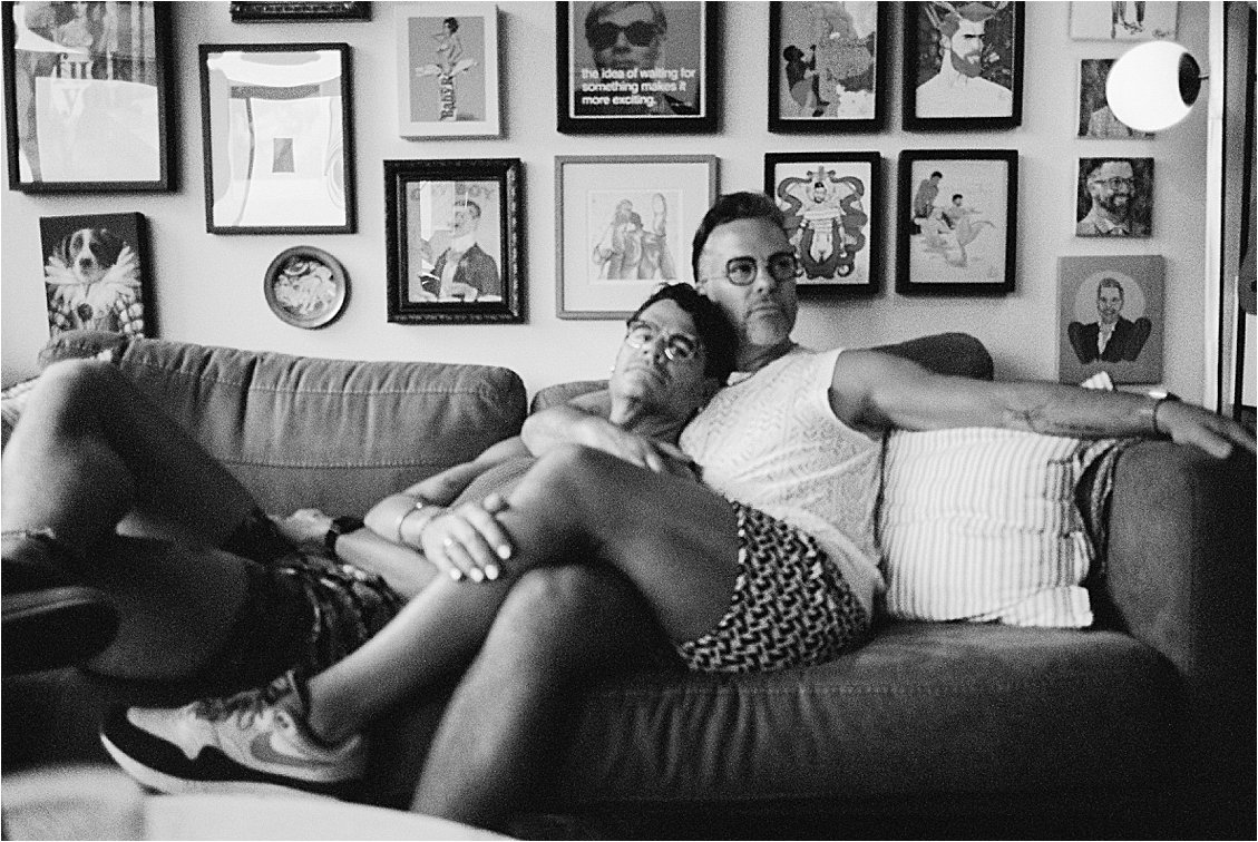 Black and white film photograph of two men lounging on couch with gallery wall behind them, Washington DC anniversary portrait session, Nikon F5