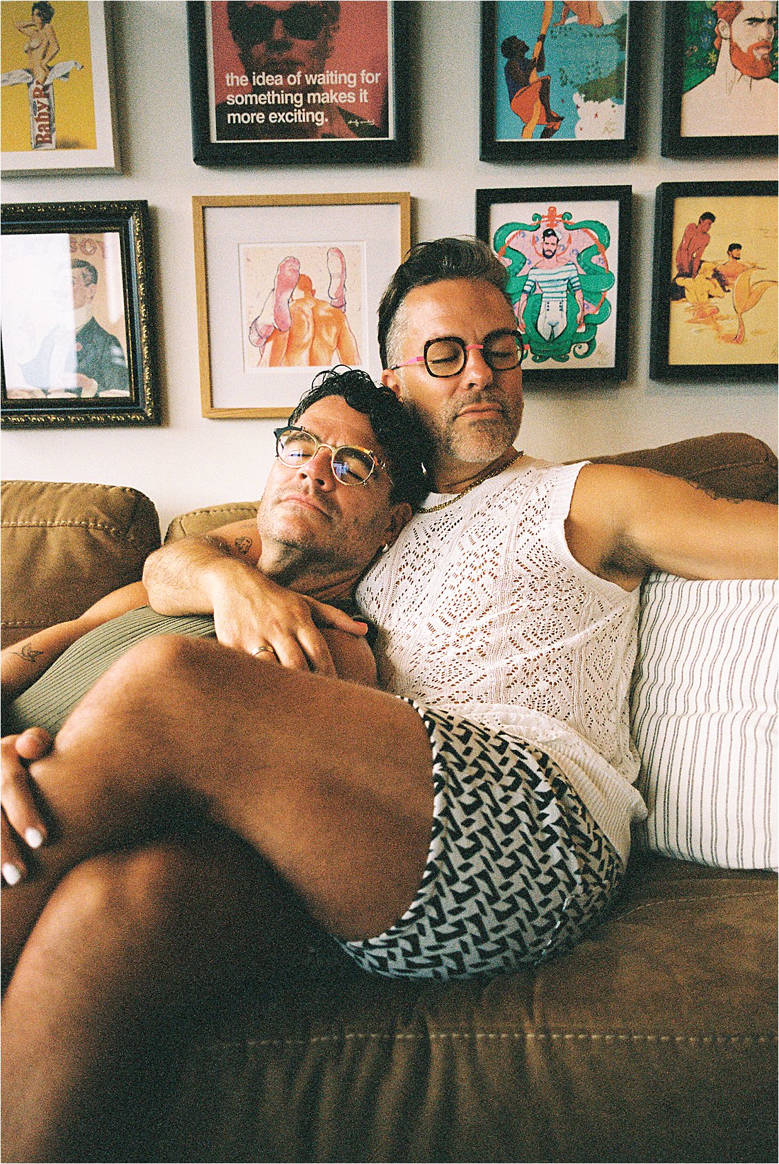 Couple portrait on couch — one man resting head on partner's shoulder with colorful gallery wall behind them, Washington DC film portrait photographer Renee Hollingshead