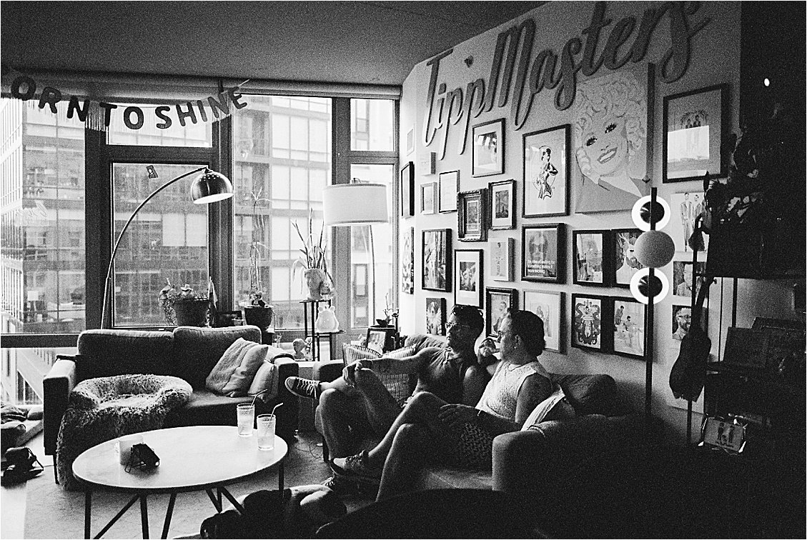 Black and white wide shot of Washington DC apartment with gallery wall, Born to Shine banner, and two men on sofa with floor-to-ceiling windows, Nikon F5 film photography