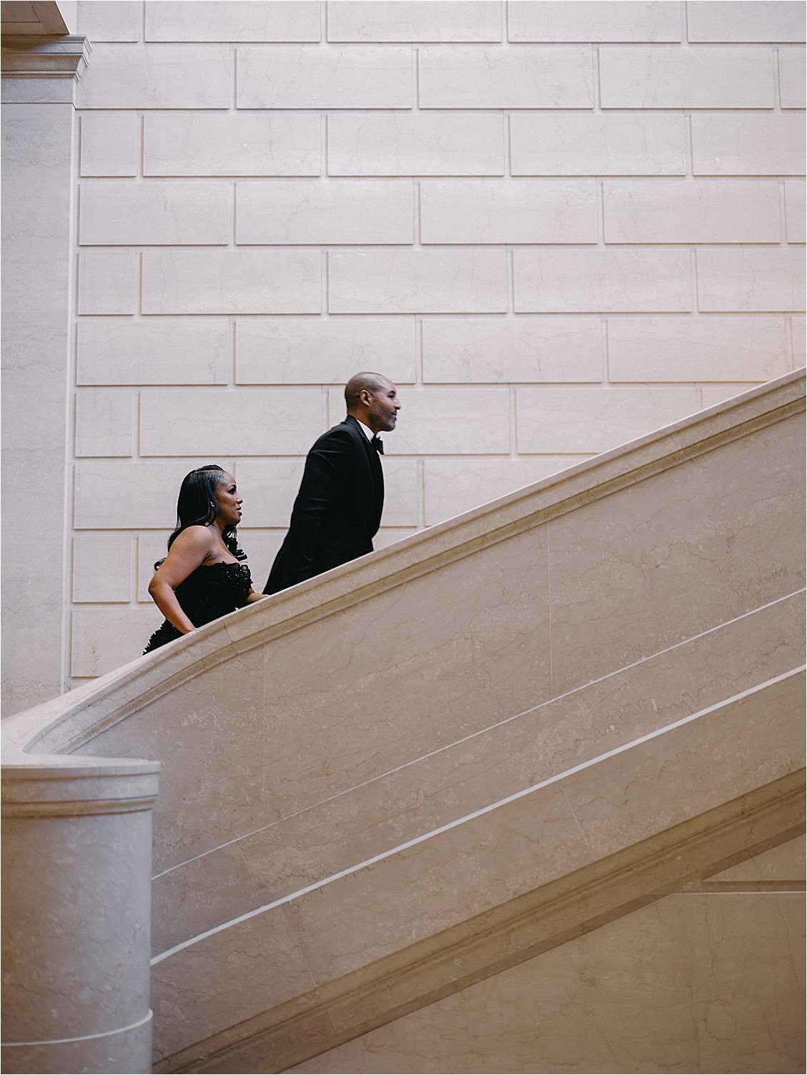 Engaged couple in black tie formalwear walking down a curved marble staircase during their National Gallery of Art engagement session in Washington, DC. Photo by destination film wedding photographer Renee Hollingshead.