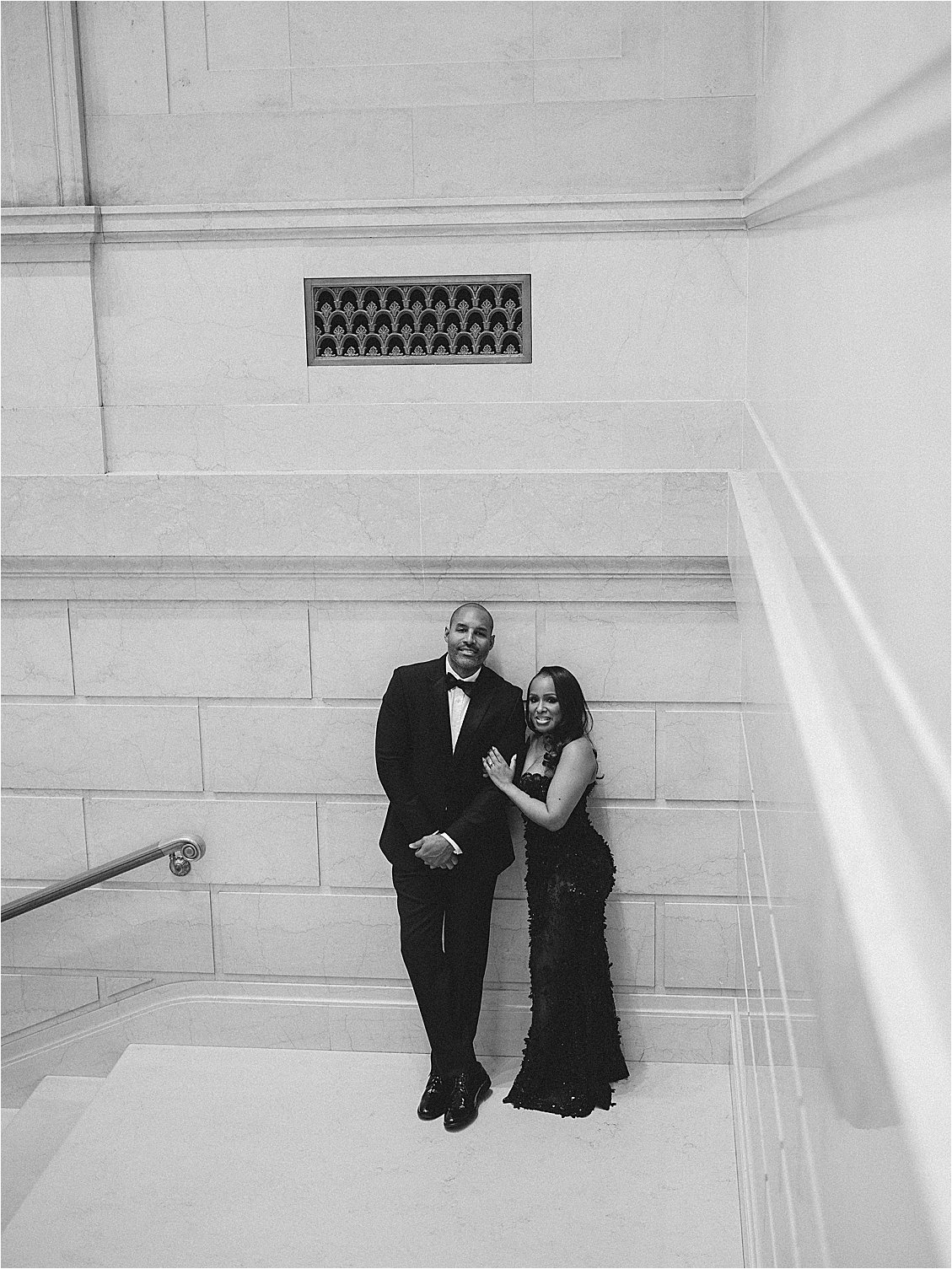 Black and white photo of an engaged couple standing together beneath an ornate decorative vent on a marble stairwell wall. Photo by destination film wedding photographer Renee Hollingshead.