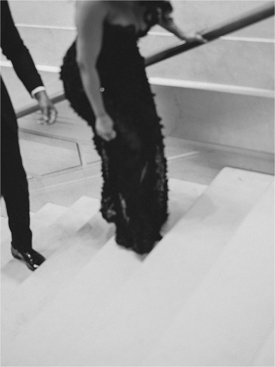 Black and white detail of a bride-to-be's black sequin gown train trailing down a marble staircase as her partner walks ahead. Photo by destination film wedding photographer Renee Hollingshead.