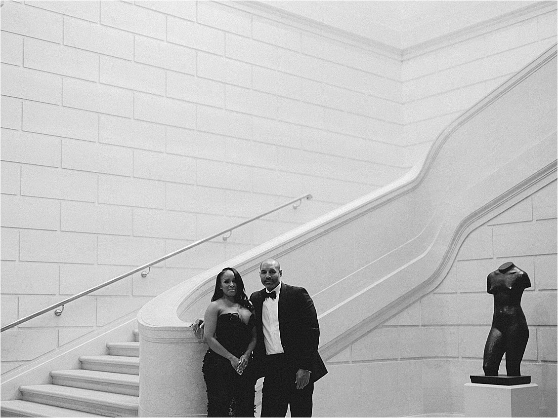 Black and white wide shot of an engaged couple standing at the base of a curved marble staircase beside a bronze statue. Photo by destination film wedding photographer Renee Hollingshead.