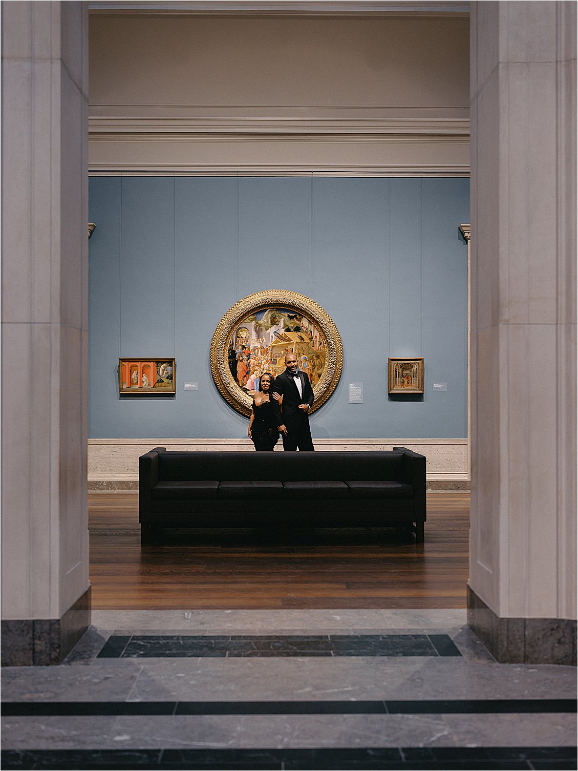 Engaged couple framed by a marble doorway standing before a gold-framed circular painting on a blue gallery wall. Photo by destination film wedding photographer Renee Hollingshead.