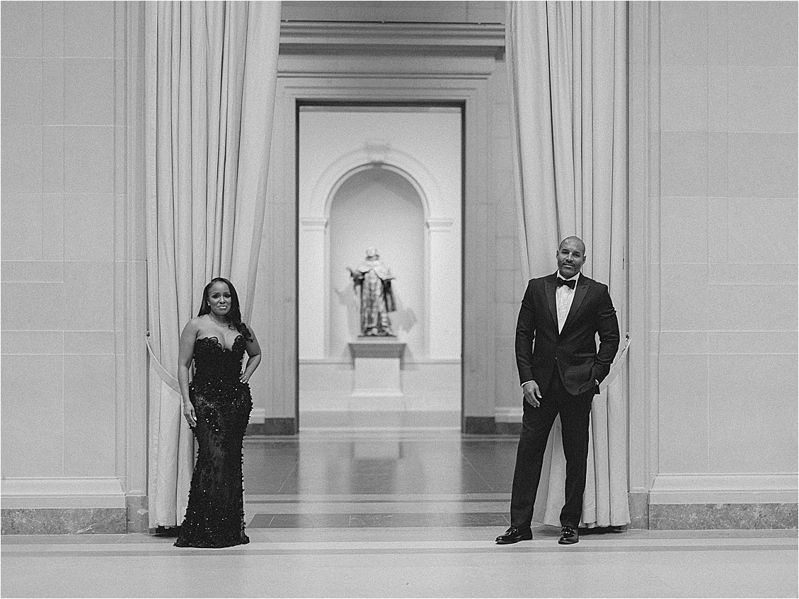 Black and white symmetrical portrait from a DC engagement session, with the couple framed beside tall draped curtains and a statue. Photo by destination film wedding photographer Renee Hollingshead.