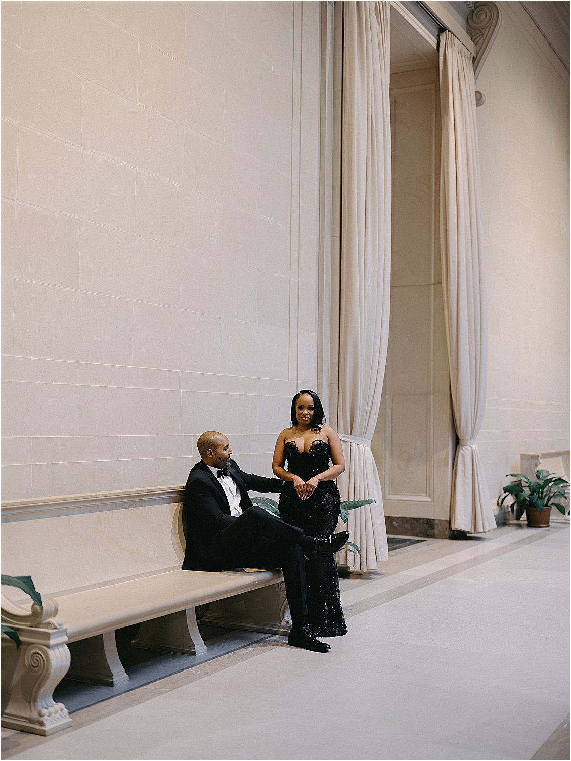 Engaged couple seated and standing beside a marble bench in a columned museum hallway lined with draped curtains. Photo by destination film wedding photographer Renee Hollingshead.
