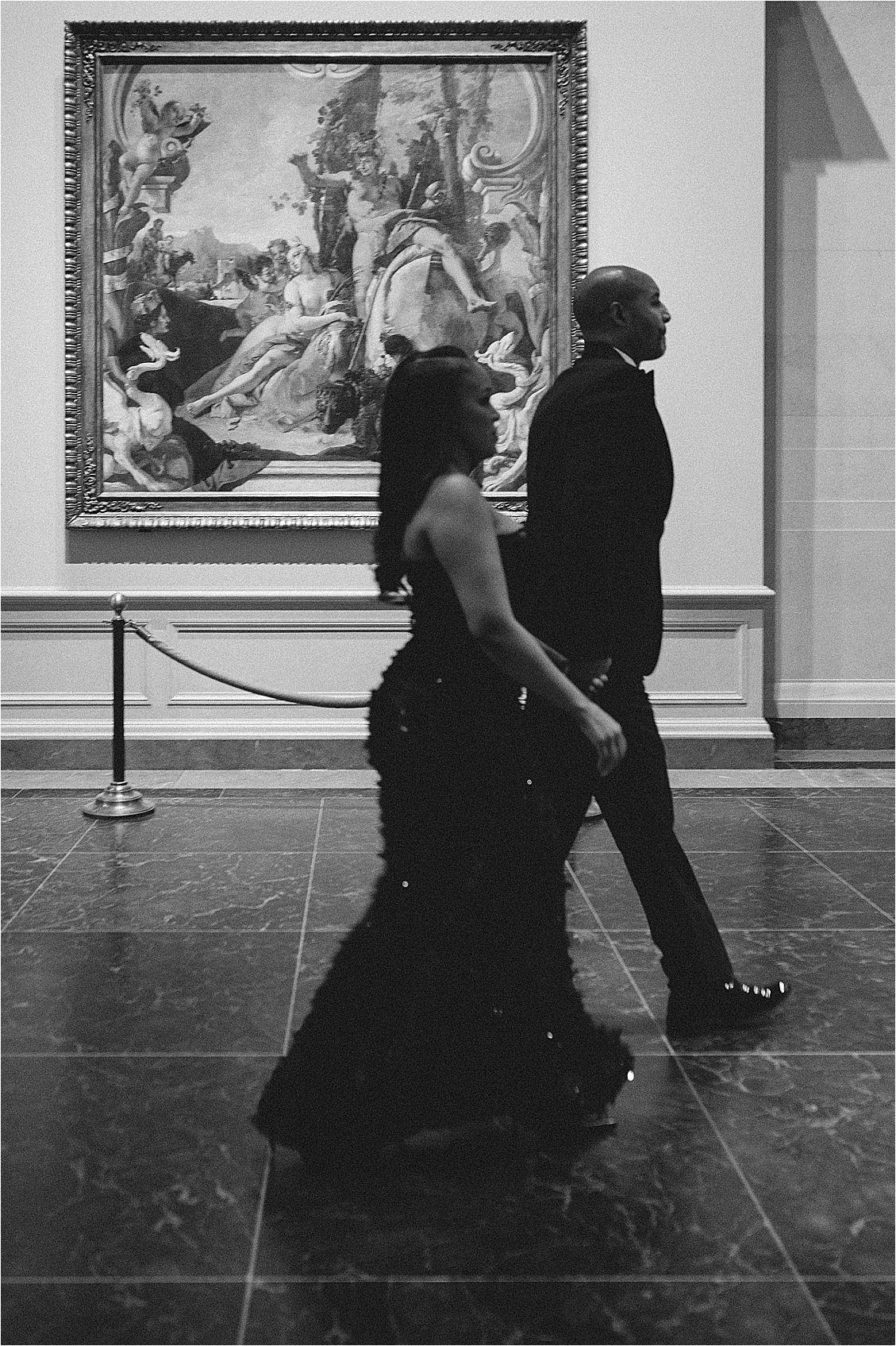 Black and white candid of an engaged couple walking past a large classical painting in a museum gallery. Photo by destination film wedding photographer Renee Hollingshead.