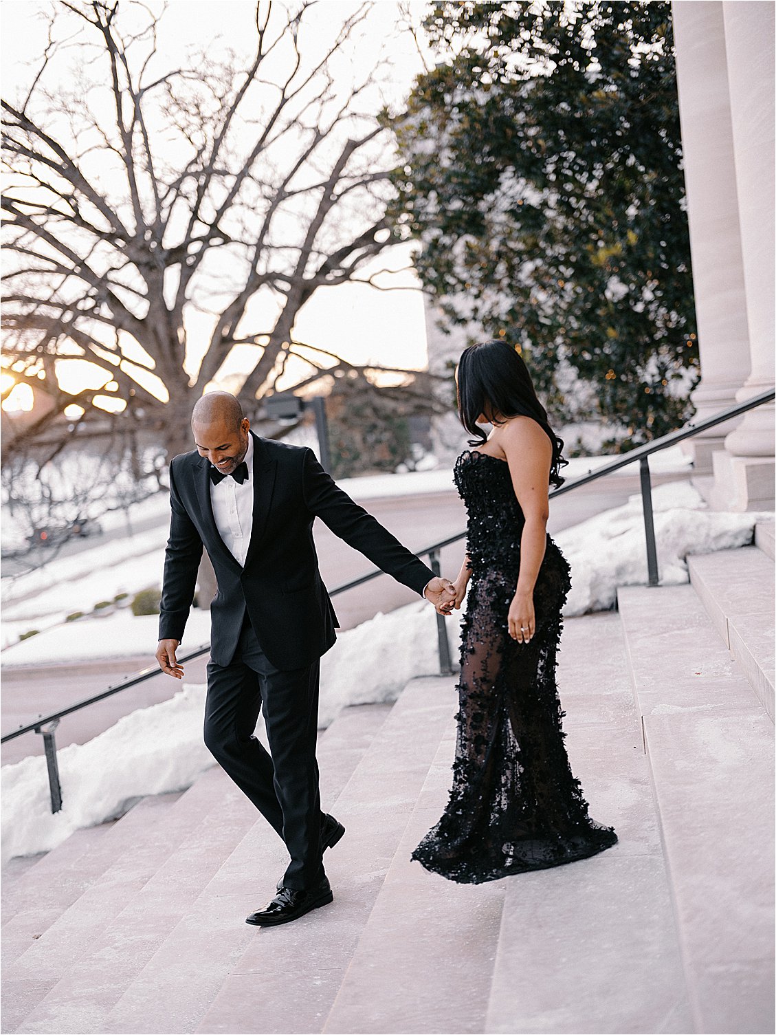 Engaged couple in black tie formalwear walking down snow-lined marble steps outside a museum at sunset. Photo by destination film wedding photographer Renee Hollingshead.